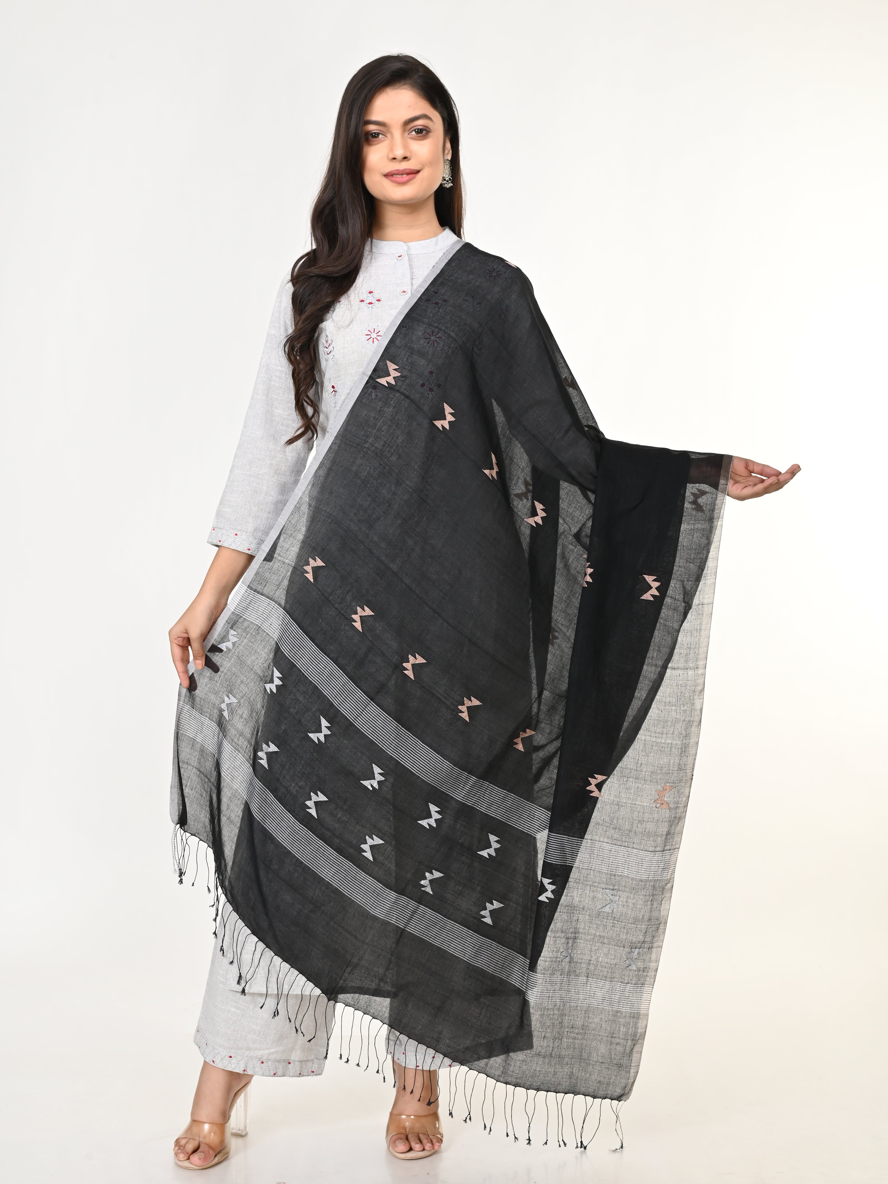 A woman modeling a black and white handloom jamdani cotton dupatta with intricate patterns and pompoms, wearing a white shirt and light-colored pants.
